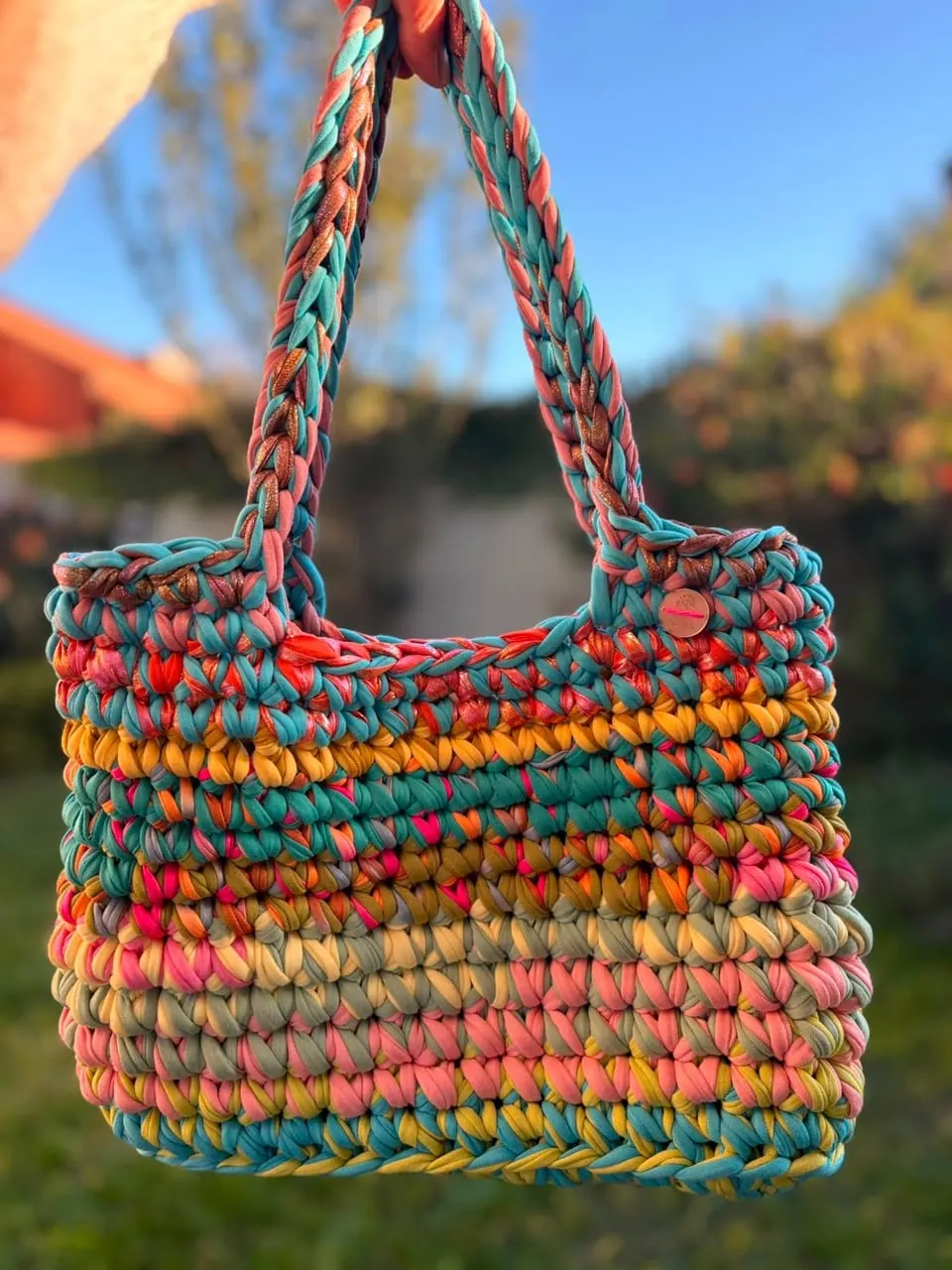 Bolso Multiuso tejido en trapillo multicolor con asas largas, fotografiado al aire libre.
